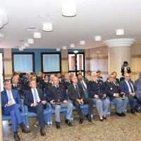 Gruppo di uomini seduti in sala conferenze, abbigliati in abiti formali, in attesa di inizio evento.