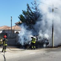 Auto avvolta dalle fiamme in un cortile urbano, vigili del fuoco all’opera con telline e manicotti, fumo grigiastro e cielo sereno.