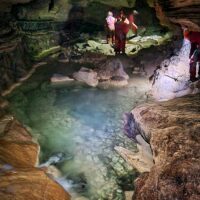 Cascata carsica limpida in una grotta: rocce, acque turchesi e due visitatori esplorano l'ambiente.