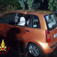 Auto arancione a 5 porte parcheggiata su prato, finestrini anteriori abbassati, alberi e notte sullo sfondo.