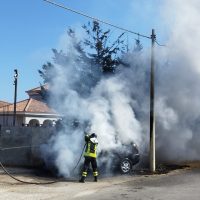 Vigile del fuoco vicino a un veicolo in fiamme, fumo grosso che avvolge l’area, scenario urbano con case e linea elettrica.