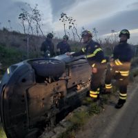 Auto incidentata capovolta su bordo strada, vigili del fuoco al lavoro in abiti riflettenti durante alba, contesto rurale.