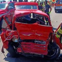 Auto rossa gravemente danneggiata in incidente, vetri rotti e frontale schiacciato, soccorritori in divisa fluorescenti all’opera.