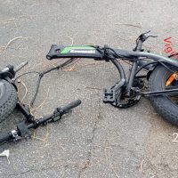 Parti di una bici elettrica smontata su strada, con ruote, manubrio e telaio visibili: componenti e cablaggi sparsi su superficie asfaltata.
