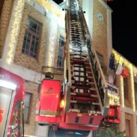 Ponteggio e scala aerea rossa vicino a edificio giallo, illuminato di notte, pompiere in azione sul veicolo antincendio.
