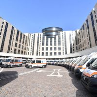 Veduta di un ospedale moderno a forma di ferro di cavallo, con facciate chiare e veicoli ambulanze parcheggiati nell’area antistante.
