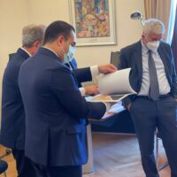 Uomini d’affari in sala conferenze, due stringono documenti; tutti indossano mascherine, atmosfera formale e professionale.