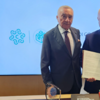 Uomo in abito scuro stringe una mano, sfondo azzurro con loghi aziendali e una persona a fianco, stemma o premio visibile sul tavolo.