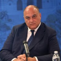 Uomo in abito scuro seduto a tavola, con microfono davanti, sfondo blå intenso e bicchiere d'acqua a destra.