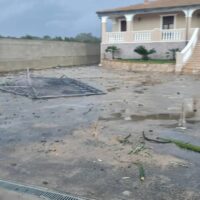 Aula al primo piano di una casa su strada, cortile sporco, pali e detriti, recinto basso e pareti rovinate, segnali di danni da alluvione.