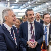 Gruppo di uomini in abiti formali in una fiera, sorridono vicino a vini in esposizione.