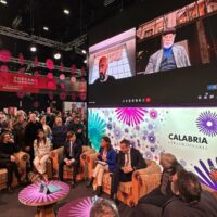 Concerto evento in sala conferenze con pubblico in cerimonia, schermi giganti mostrano relatori, decorazioni viola e logo Calabria sullo sfondo.