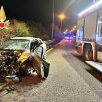 Incidente stradale notturno: auto danneggiata e scooter a terra, soccorsi in corso con camion di soccorso, luce neon e strada bagnata.