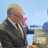 Due uomini in abiti formali si scambiano una brochure o un libro, sullo sfondo uno schermo con logo 'adnkron'.