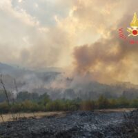 Paesaggio di primo piano con fumo e braci diffuse, cielo illuminato dal tramonto, elementi di vegetazione e terreno secco.