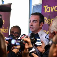 Uomo politico al centro, intervistato da diverse televisioni, circondato da giornalisti e crowd in primo piano.