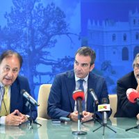 Tre uomini in un tavolo da conferenza, con microfoni, in posa durante una riunione; colori vividi sullo sfondo blu.