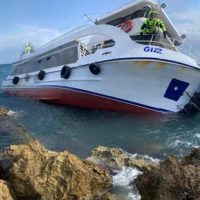 Barca a motore bianca ormeggiata tra rocce coste, vista laterale, equipaggio sul ponte, mare sullo sfondo, cielo sereno.