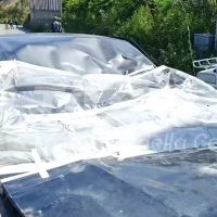 Auto danneggiata coperta da teloni trasparenti, contesto urbano con alberi e veicoli sullo sfondo.