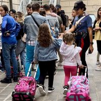 Gruppo di studenti in fila con trolley colorati, maestra al centro e bambini che aspettano, in una scena di scuola o attività educativa.