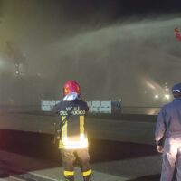 Operai al lavoro in una situazione notturna, indossano giubbotti riflettenti e caschi, luce di emergenza e fumo visibile sullo sfondo.