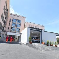 Azienda moderna con edificio beige e grandi finestre: ingresso principale affollato da due persone, zona uffici e area esterna con auto parcheggiate.