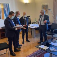 Quattro uomini in abiti formali discutono documenti su un tavolo, in una stanza d'ufficio con logo e dipinti alle pareti.