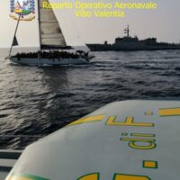 Barca a vela in primo piano, mare calmo e yacht in lontananza; velivoli operativi e testo in alto, scena marina italiana.