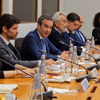 Foto di un gruppo di uomini in riunione, seduti al tavolo con documenti, bottiglie d'acqua e microfoni, intento a discutere.