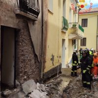 Cornice crollata e detriti lungo una strada stretta; vigili del fuoco e soccorritori in divise nere e arancioni a bordo, tra edifici fatiscenti.