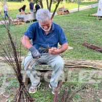 Uomo anziano al lavoro, intento a intrecciare rami secchi in un giardino pubblico, sfondo prato e persone distanti.