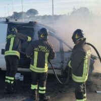 Tre vigili del fuoco in divisa riflettente insieme a un veicolo incendiato, fumo visibile e intervento in corso su strada.