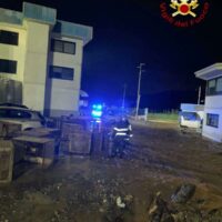 Strada notturna con veicoli fermati e luci lampeggianti, edifici residenziali sullo sfondo e terreno irregolare in primo piano.