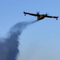 Aereo a turboelica in volo spezza la fascia di nuvole con scia di fumo sul cielo azzurro.