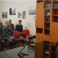 Sala conferenze con ufficiali in uniforme seduti su sedie rosse, scaffali in legno e foto nere bianche alle pareti.