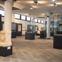 Interno di museo luminoso con vetrine e display, colonne bianche, pavimento in pietra e luci soffuse, atmosfera tranquilla.