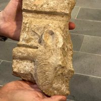 Schema in pietra con incisioni ad andamento astratto; mani che lo sostengono, fondo di gradini in pietra.