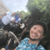 Viaggiatore felice all’aperto con cappellino, bordo boscoso e amici sullo sfondo: selfie racconta momenti di avventura e natura.