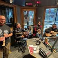 Gruppo di amici in studio radiofonico, seduti intorno a tavolo con microfoni, monitor e registratori, atmosfera conviviale e sorridente.