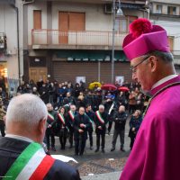 Cattura una cerimonia pubblica all'aperto: un vescovo in abiti viola benvenuti, un uomo con fascia tricolore e folla dietro di lui.