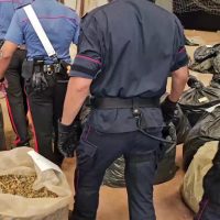 Due ufficiali in uniforme osservano una scena con sacchi e cavi, sullo sfondo macchinari e altri oggetti; l’immagine è in bocca d’uso.