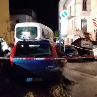 Auto parcheggiate in una via serale, con una barriera rossa di cantiere e un vicolo stretto tra edifici.