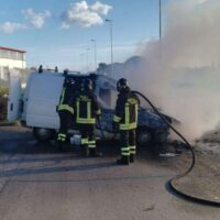 Operatori antincendio in mezzo a fumo denso proveniente da un veicolo di soccorso, erba allontanata, maniche e attrezzature a terra.