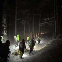 Escursionisti in un bosco innevato di notte, illuminati da torce e lampioni, camminano lungo un sentiero tra alberi scuri.