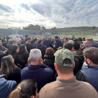 Grandi persone si riuniscono all'aperto in gruppo, in una giornata nuvolosa vicino a una recinzione, probabilmente in un evento pubblico.