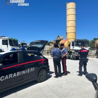 Gruppo di Carabinieri intervengono vicino a veicoli e una grande torre cilindrica gialla in un'area all'aperto, cielo sereno.