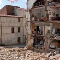Edificio parzialmente crollato con detriti, persone tra pavimenti sbriciolati e architettura deserta, cielo luminoso.