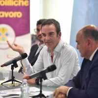 Uomo in camicia bianca parla al convegno, seduti a tavolo con microfoni, altri relatori sullo sfondo e banner colorato.