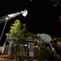 Immagine notturna: gru telescopica e nastro rosso fermano un cantiere, luci artificiali illuminano la scena davanti a un edificio e alberi.