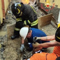 Operai in cantiere indossano caschi: due manodopera collaborano per scavo tra intonaco e terreno, con equipaggiamento di protezione.